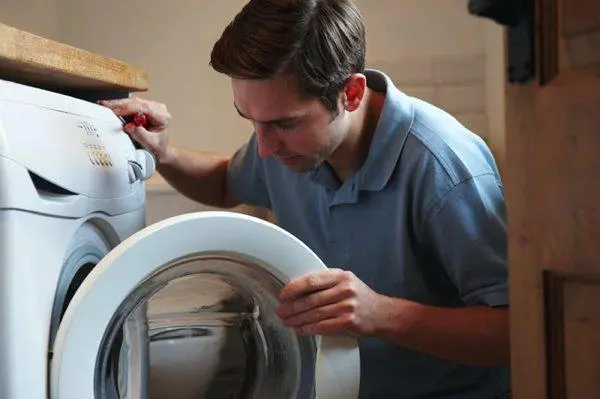 A professional male appliance technician in a blue polo shirt inspecting a white front-loading washing machine in a home laundry area.